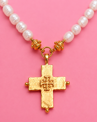 Gold cross pendant on a pearl necklace against a pink background