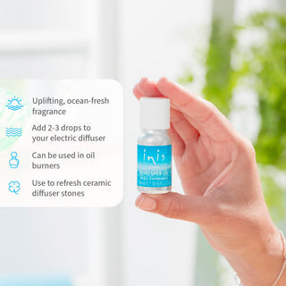 Hand holding a small bottle of 'inis' refresher oil with product details on a blurred natural background