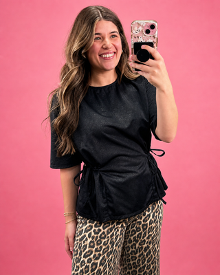 Woman taking a selfie with a phone against a pink background, wearing a black top and leopard print pants.
