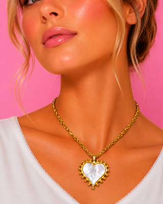 Close-up of a person wearing a gold necklace with a heart-shaped pendant against a pink background