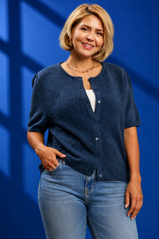 A woman with blonde hair wearing a dark blue short-sleeve button-up cardigan over a white top and light blue jeans, standing against a blue background with windowpane shadows.