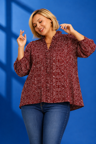A woman modeling a long-sleeve button-up blouse with a dense small-scale red, burgundy, and white speckled pattern, paired with blue jeans against a vibrant blue studio background.