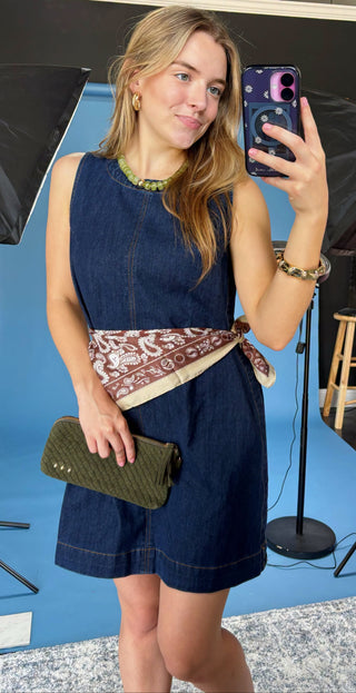 A woman takes a mirror selfie wearing a sleeveless dark blue denim romper with a brown and beige patterned sash tied at the waist, holding an olive green clutch. The background shows a studio setup with blue backdrop paper, softbox lights, and a stool.
