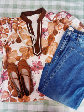 A styled flat-lay outfit featuring a cream-colored short-sleeve top with a brown and pink floral pattern and a brown V-neckline, paired with blue jeans, brown woven flat shoes, and a white beaded necklace