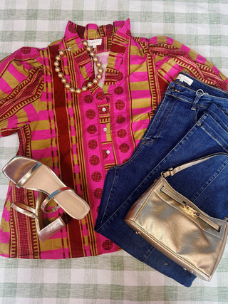 A styled flat-lay outfit on a green and white checkered fabric background, featuring a vibrant pink and gold patterned short-sleeve blouse, dark blue jeans, a gold metallic shoulder bag