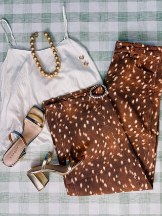 A styled flat-lay outfit featuring brown and white spotted wide-leg denim shorts, a white spaghetti strap camisole, a gold beaded necklace, and metallic gold block-heel sandals, all arranged on a green and white checkered blanket.