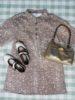 A short-sleeve collared shirt dress in taupe with a white speckled pattern, styled in a flat lay with white Mary Jane shoes and a metallic gold handbag on a green and white checkered blanket.