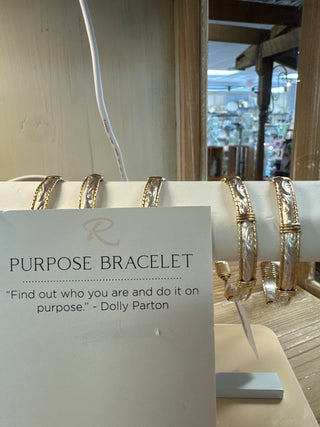 Gold bracelets on a white stand with a sign about 'Purpose Bracelet' in a store setting.