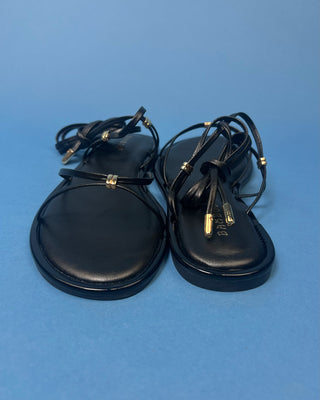 A pair of black leather flat sandals with thin strappy details and small gold hardware, against a solid blue background.