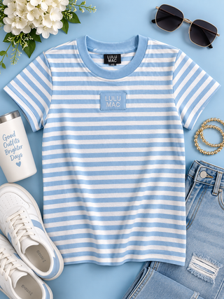A light blue and white striped short-sleeve t-shirt with a small square patch reading 'LULU MAC' on the chest, styled in a flat lay with sunglasses, jeans, sneakers, a tumbler, and flowers on a light blue background.