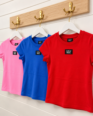 Three children's short-sleeve T-shirts in pink, blue, and red, hanging on white hangers from gold wall hooks on a wooden board against a white paneled wall. Each shirt has a small black chest label reading 'LULU MAC'.
