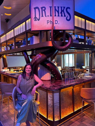 A woman in a green and white striped dress sits on a bar stool in a modern bar setting with a large octopus sculpture and a hanging sign reading 'DR.INKS Ph.