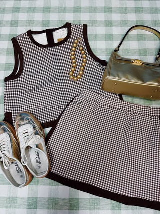A coordinated outfit laid flat on a green-and-white checkered surface, featuring a sleeveless black-and-white gingham top with dark trim, a matching gingham skirt, a gold-tone handbag, a beaded necklace, and white sneakers.