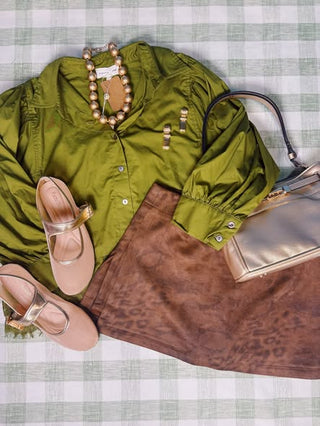 A styled flat-lay outfit on a green and white checkered blanket, featuring an olive green long-sleeve button-up shirt, a brown leopard-print skirt, beige Mary Jane shoes, a gold beaded necklace, and a metallic gold handbag.
