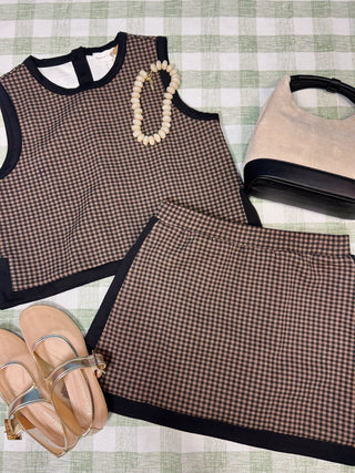 A coordinated outfit laid flat on a green-and-white checkered surface, featuring a brown and beige gingham sleeveless top with black trim, a matching gingham mini skirt, beige flat sandals, a cream beaded necklace, and a beige handbag with black trim.