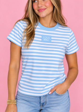A woman wearing a light blue and white striped t-shirt with a small 'LULU MAC' logo on the chest, paired with light blue jeans, set against a solid pink background.
