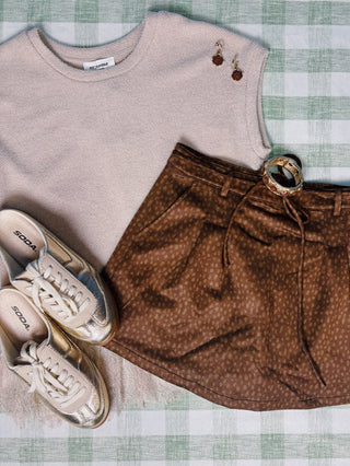 A styled flat-lay outfit featuring a light beige knit sleeveless top, brown patterned shorts with a round metal buckle belt, beige and silver sneakers, and brown dangling earrings, all arranged on a green and white checkered blanket.