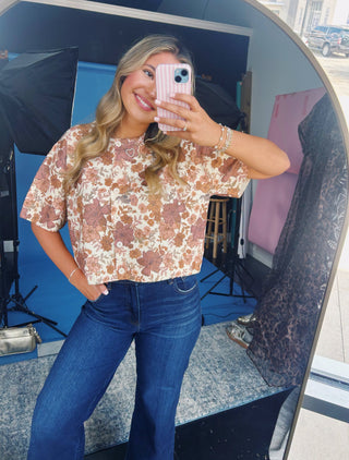 A woman takes a mirror selfie wearing a short-sleeve floral top with blue jeans. The top has a cream base with brown and muted orange floral patterns.
