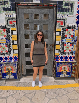 A woman wearing a sleeveless ribbed gray mini dress with black trim, white sneakers, and sunglasses stands in front of a rustic wooden door set within a colorful mosaic tile wall.