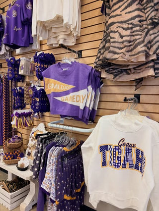 A retail display of purple and white custom apparel, featuring a purple cropped t-shirt with 'GAMEDAY GAMEDY' text, a white sweatshirt with 'Gears TUGAR' in blue and gold varsity lettering, and other matching garments hanging on racks against a wood slatwall.