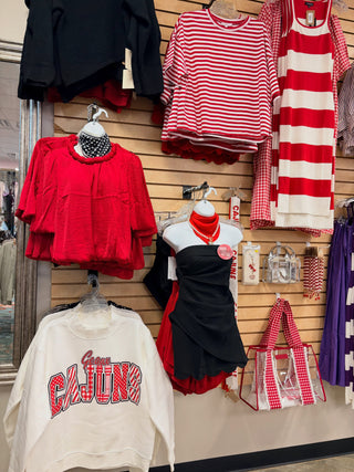 A retail display of various women's clothing items hanging on a slatwall, including red and white striped tops, a red tiered blouse, a black strapless dress, a white sweatshirt with 'Cajuns' text, and patterned handbags.