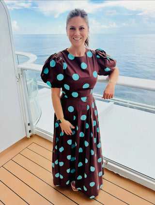 A woman stands on a cruise ship balcony wearing a brown maxi dress with large turquoise polka dots. The background shows the ocean and sky, with the wooden deck visible beneath her.