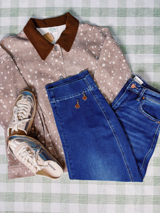 A styled flat-lay outfit featuring a beige long-sleeve button-up shirt with white speckles and a brown corduroy collar, paired with blue jeans and metallic silver sneakers on a green and white checkered blanket.