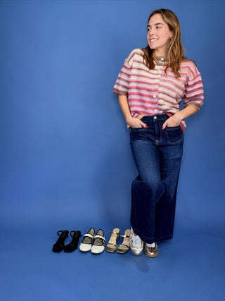 A woman stands against a solid blue studio background wearing a pink and white striped knit top, dark blue wide-leg jeans, and metallic sneakers. A row of six shoes in different colors and styles is lined up on the floor to her left.