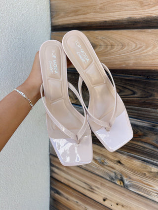 A pair of nude pink patent leather thong sandals with square toes and low block heels, held by a hand against a background of a textured white wall and rustic wooden planks. 