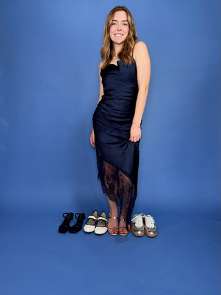 A woman stands against a blue studio backdrop wearing a dark navy satin midi dress with sheer black lace tights. Four pairs of shoes are placed on the floor in front of her
