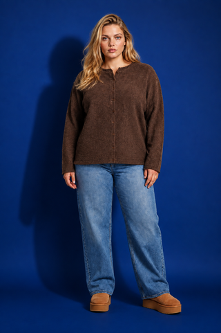 A woman modeling a brown fuzzy long-sleeve button-front cardigan with blue jeans and tan slip-on shoes, standing against a solid blue studio background.
