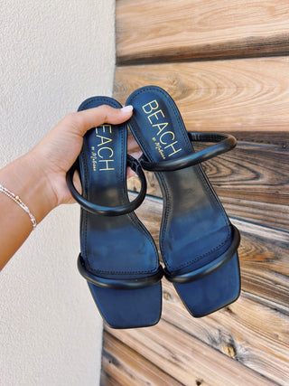 A hand holds a pair of navy blue leather slide sandals with a single thick strap and square toes