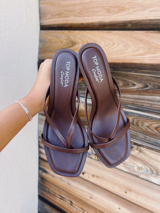 A pair of brown square-toe thong sandals with a low block heel, held by a hand against a rustic wooden plank background. 