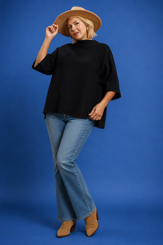 A blonde woman wearing a black oversized mock-neck top and light blue denim shorts, posing against a solid blue studio background.