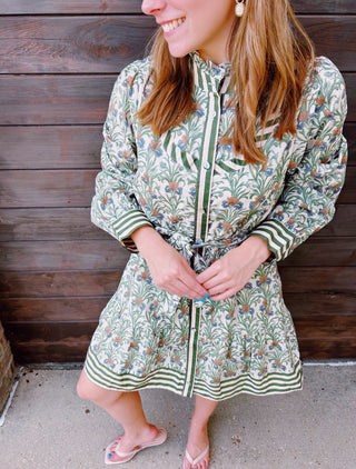 A woman wearing a long-sleeve button-front dress with a green, white, and orange botanical pattern, standing in front of a dark wooden plank wall.