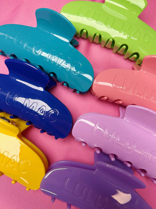 A set of colorful Lulu Mac brand hair claw clips arranged on a solid pink background. The clips are shown in multiple vibrant colors including teal, lime green, navy blue, pink, yellow, lavender, and purple.