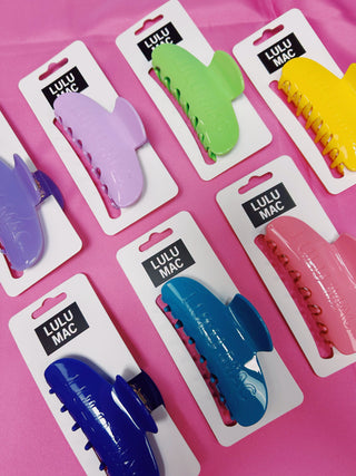 Six colorful hair claw clips in purple, green, yellow, pink, blue, and dark blue, each attached to a white retail card labeled 'LULU MAC' and arranged on a bright pink background.