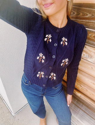 Navy cable knit cardigan with embroidered white daisies, button front, crew neckline, and long sleeves.