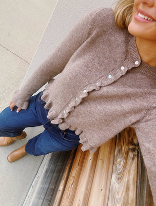 Taupe knit cardigan with delicate ruffle trim, pearl-style buttons, ruffled hem, and feminine flared cuffs.