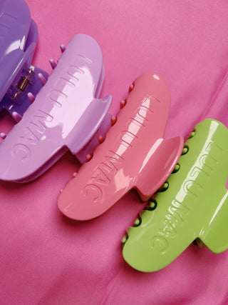 Three glossy hair claw clips in lavender, pink, and lime green, arranged diagonally on a textured pink background. The clips feature the brand name 'LULU MAC' embossed on the front.