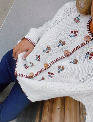 a cream cable-knit button-up sweater with embroidered floral details and brown buttons