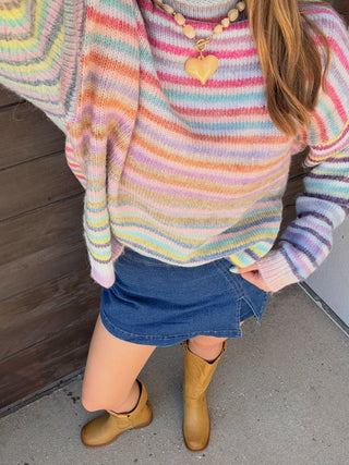 A person wearing a pastel rainbow striped knit sweater, a blue denim mini skirt, and tan cowboy boots, standing outdoors against a dark wooden door and concrete ground.
