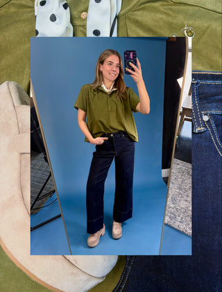 A woman takes a mirror selfie wearing an olive green short-sleeve top and dark navy wide-leg pants, set against a blue studio background. The photo is layered over a collage of clothing textures, including denim, olive fabric, and a polka-dot shirt.