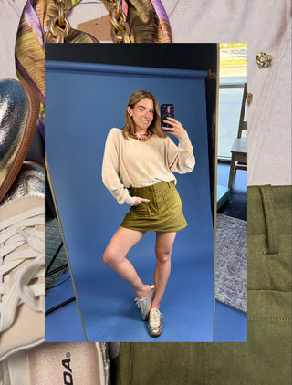A woman takes a mirror selfie wearing a beige long-sleeve knit top and an olive green mini skirt, set against a blue studio backdrop. The image is framed by a collage of other fashion items, including a metallic handbag, a white shirt, and a pair of white sneakers.