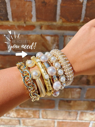 A stack of diverse gold and pearl bracelets worn on a person's wrist, set against a rustic brick wall background with a text overlay reading 'BRACELETS you need!'.