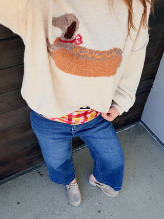 A person wearing a cream knit sweater with a duck motif, blue wide-leg jeans, and gray slip-on shoes, standing against a dark wooden wall.