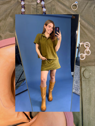 A woman takes a mirror selfie wearing an olive green short-sleeve top and matching mini skirt set, paired with tan knee-high boots, against a blue studio backdrop. The image is layered over a collage of olive green clothing and jewelry.