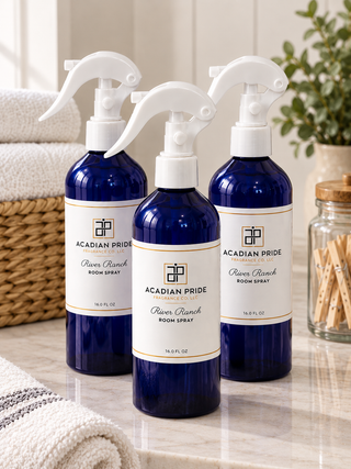 Three cobalt blue room spray bottles with white spray tops and white labels reading ACADIAN PRIDE River Ranch ROOM SPRAY, arranged on a bathroom counter with folded towels and a plant in the background.