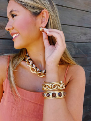 A woman wearing gold-toned jewelry, including a chunky chain necklace, stacked bracelets, and a small hoop earring, posing against a dark wooden plank background.