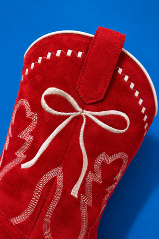 A close-up of a red suede cowboy boot against a solid blue background, featuring white embroidered details including a bo